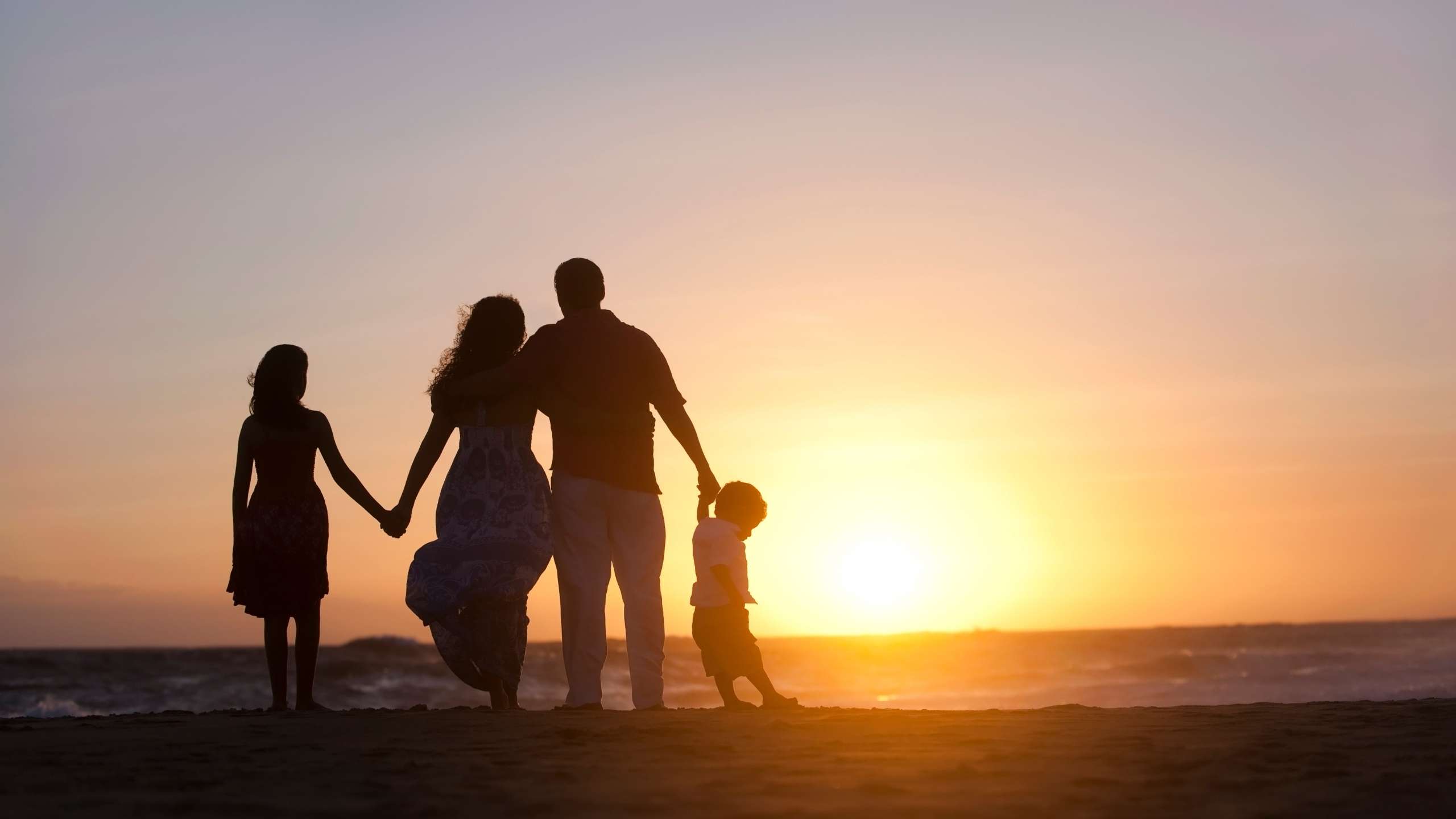 Silhouette of a family of four holding hands on a beach at sunset with warm golden light on the horizon