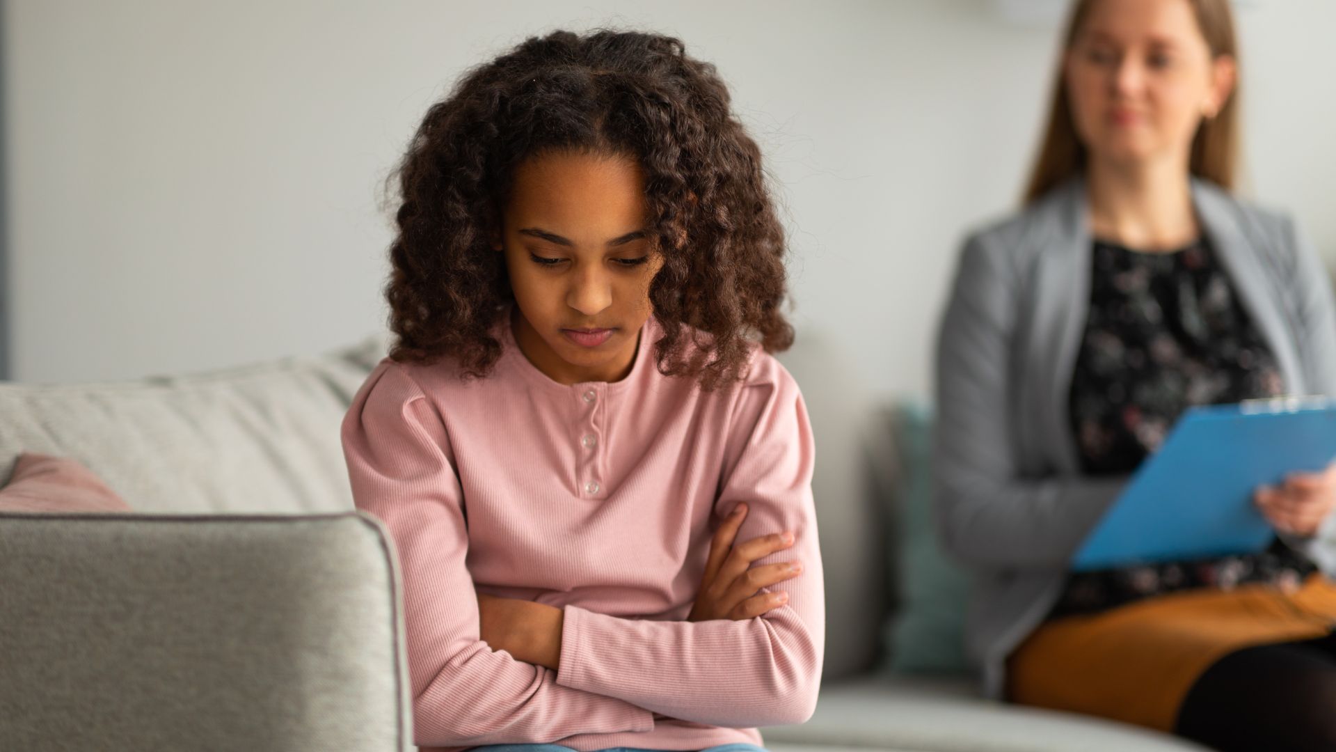 Young girl with curly hair sitting on a couch with arms crossed looking downward while a therapist holds a clipboard in the background