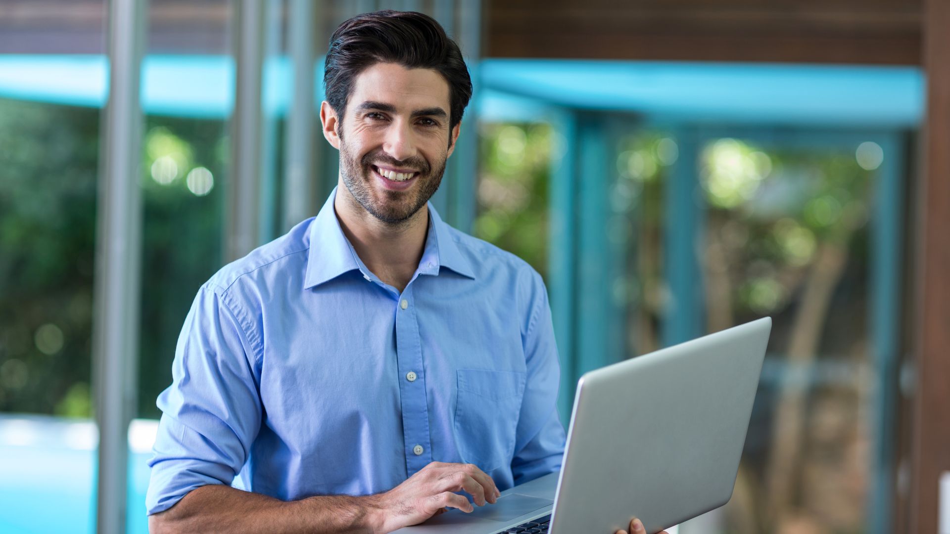 Smiling man in a blue button-up shirt working on a laptop in a bright, modern space